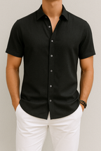Homme portant une chemise noire à manches courtes, élégante et décontractée, avec un pantalon blanc. Mode masculine, style estival chic.