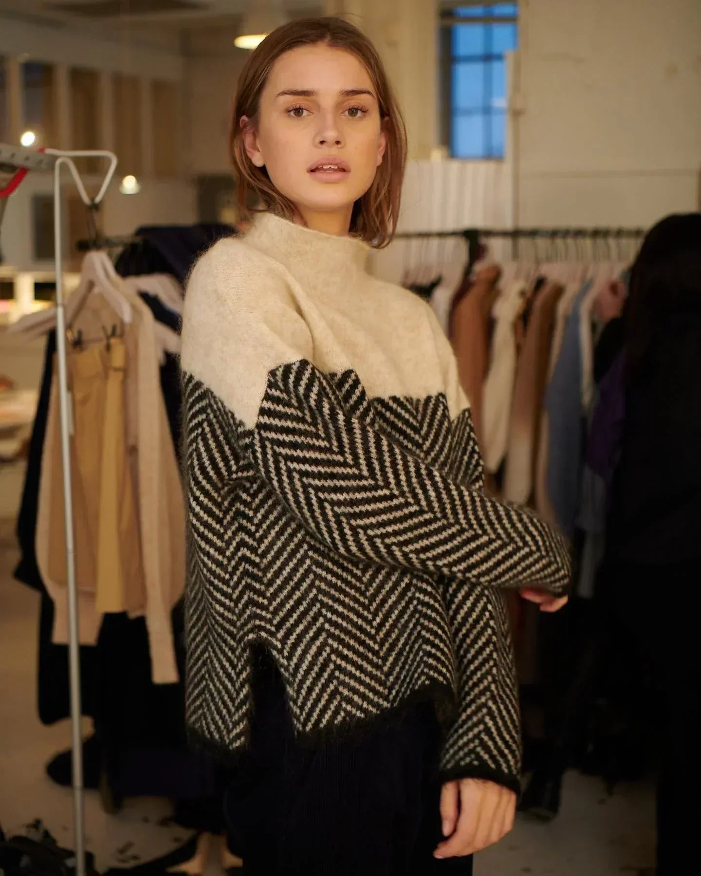 Jeune femme portant un pull en laine à motif chevron noir et blanc, posant dans un studio de mode avec des vêtements suspendus en arrière-plan.