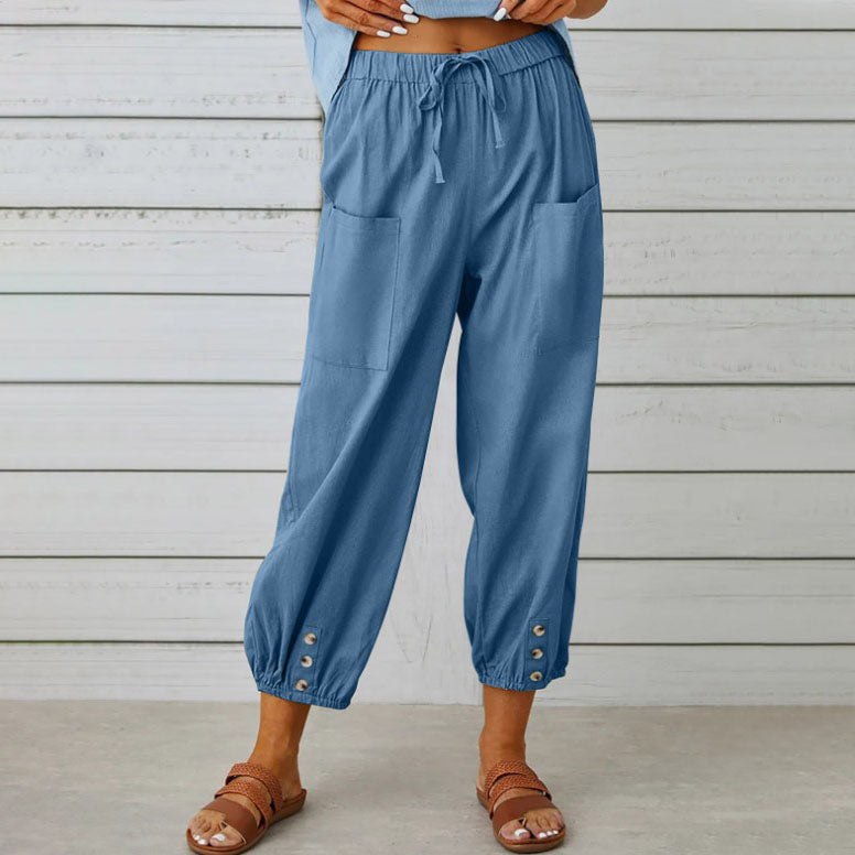 Pantalon bleu ample avec poches avant, boutons décoratifs aux chevilles, porté avec des sandales marron sur fond de mur en bois blanc.