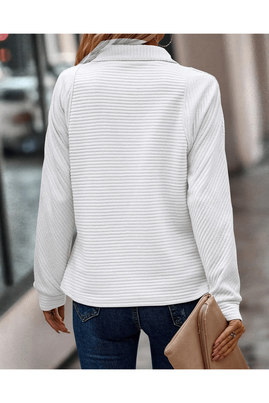Femme portant un pull blanc côtelé, manches longues, avec un sac à main beige, marchant en ville. Mode automne, style décontracté, tendance urbaine.