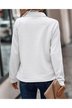Femme portant un pull blanc côtelé, manches longues, avec un sac à main beige, marchant en ville. Mode automne, style décontracté, tendance urbaine.