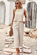 Femme en tenue beige élégante, portant un sac en osier, marchant sur un chemin de pierres. Mode estivale, style décontracté chic, accessoires dorés.