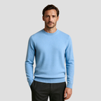 Homme portant un pull bleu clair en laine, manches longues, col rond, sur fond gris. Mode masculine élégante, tenue automne-hiver.