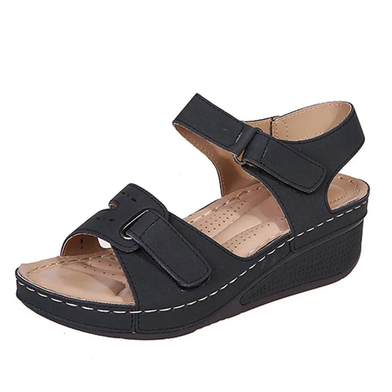 Sandale compensée noire pour femme avec brides ajustables en velcro, semelle intérieure beige et surpiqûres blanches. Chaussure confortable et élégante.