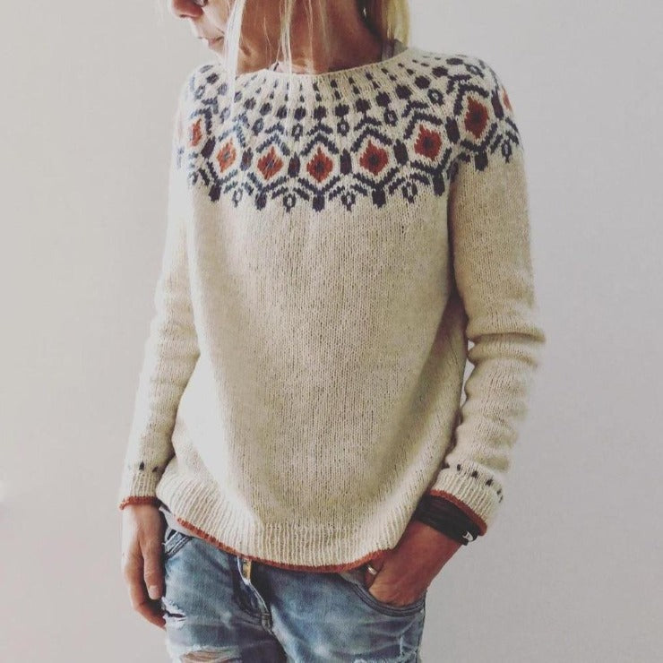 Femme portant un pull en laine beige avec motif nordique coloré, manches longues, col rond, associé à un jean décontracté. Mode hiver tendance.