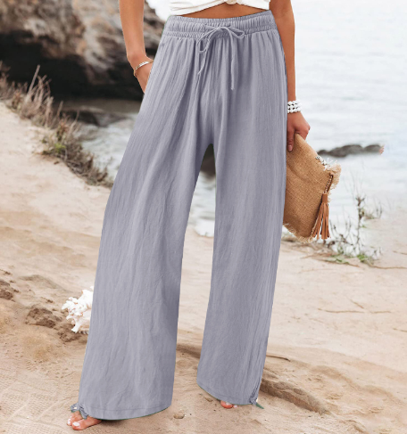 Pantalon large gris clair en lin pour femme, style décontracté, plage en arrière-plan, accessoire sac en paille, mode estivale élégante.