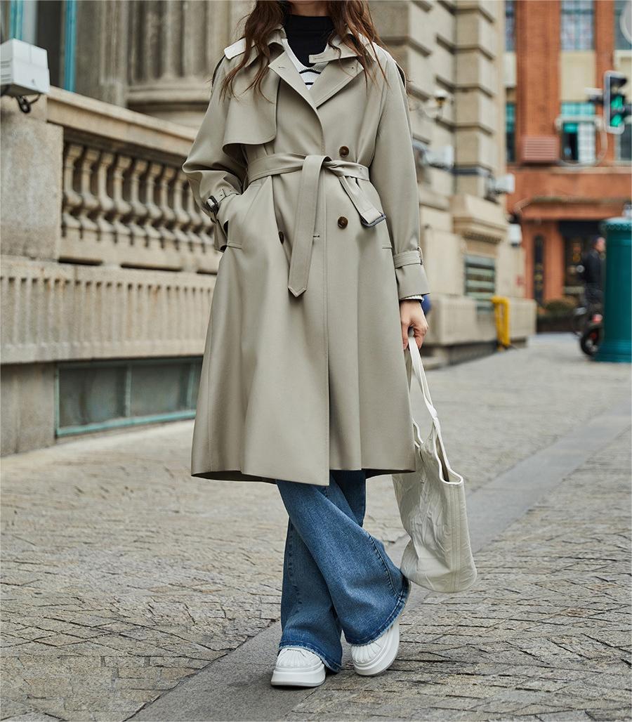 Femme en trench-coat beige, jeans évasés et baskets blanches, tenant un sac en toile, marchant en ville. Mode automne, style urbain chic.