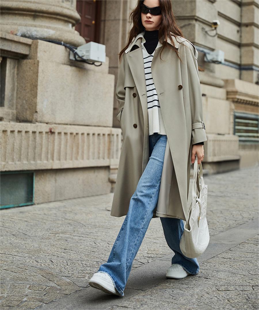 Femme portant un trench-coat beige, pull rayé, jean bleu et baskets blanches, marchant en ville avec un sac blanc. Mode urbaine chic.