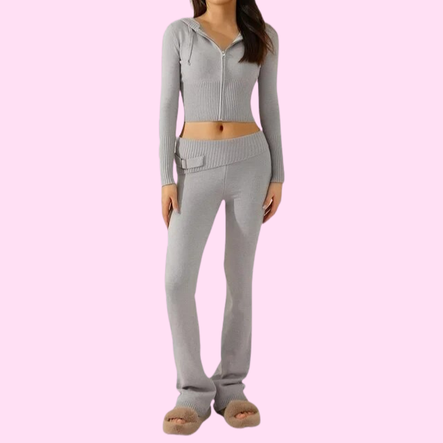 Femme portant un ensemble de survêtement gris, composé d'un haut à capuche zippé et d'un pantalon évasé, sur fond rose. Mode décontractée tendance.