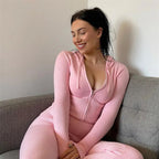 Jeune femme souriante en combinaison rose, assise sur un canapé gris. Mode décontractée, tenue confortable, ambiance détendue.