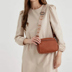 Femme en robe beige portant un sac bandoulière marron avec sangle à motifs géométriques. Mode féminine, accessoire tendance, style décontracté.