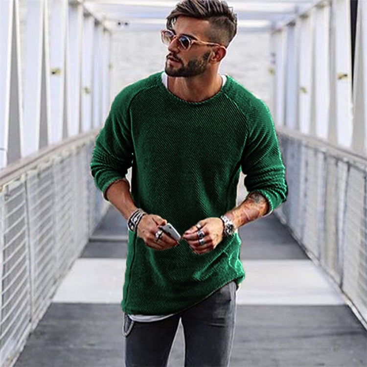 Homme portant un pull vert, lunettes de soleil et jeans, marchant sur un pont moderne. Mode masculine, style décontracté, accessoires tendance.