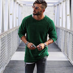 Homme portant un pull vert, lunettes de soleil et jeans, marchant sur un pont moderne. Mode masculine, style décontracté, accessoires tendance.