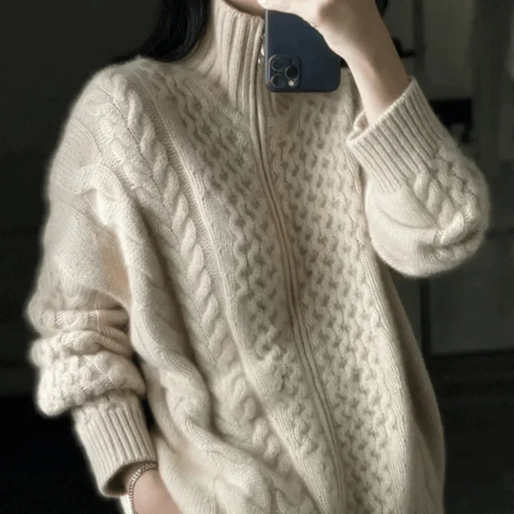 Femme portant un pull en laine torsadée beige avec fermeture éclair, tenant un smartphone. Mode automne-hiver, style confortable et élégant.