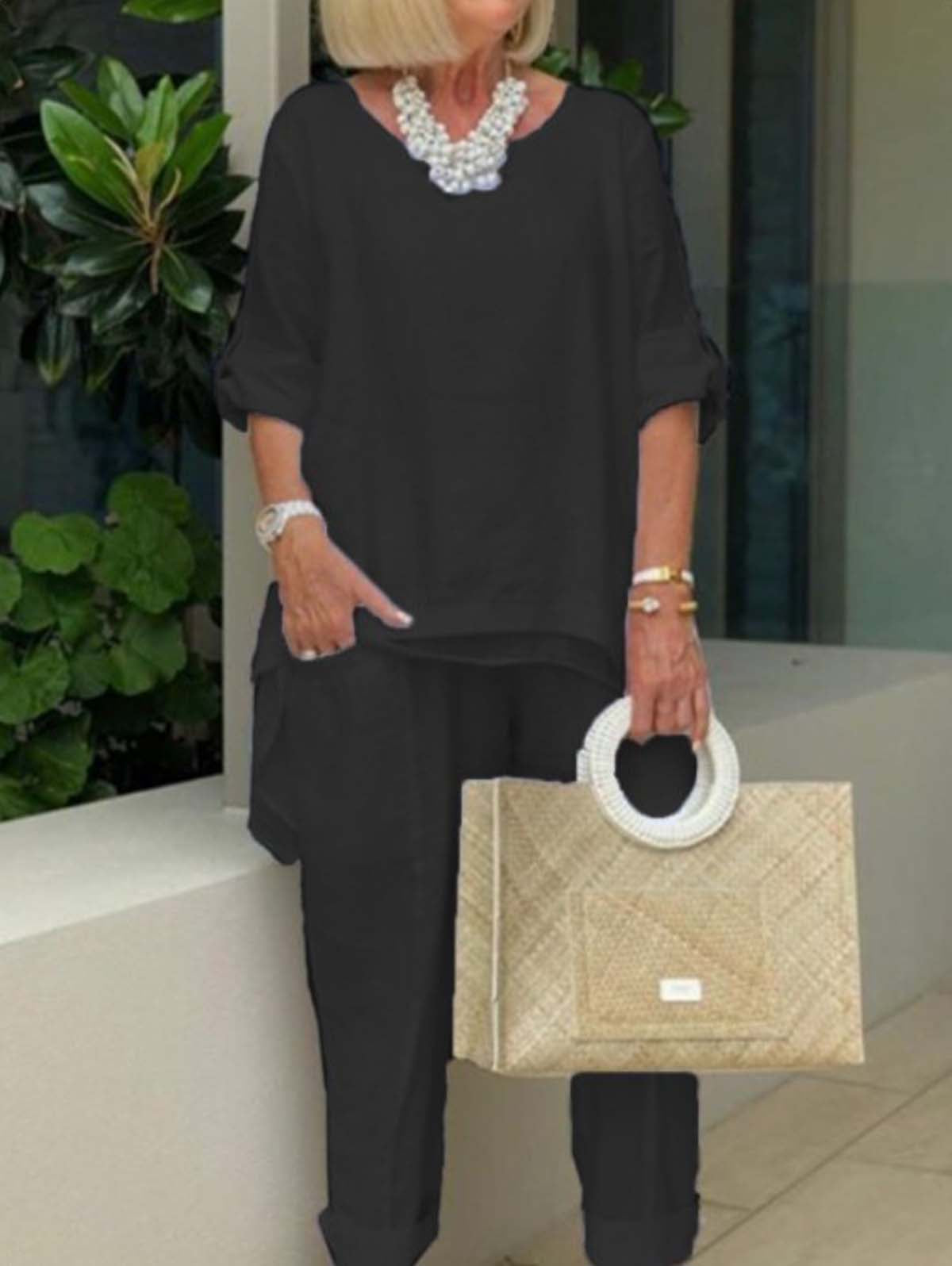 Femme élégante en tenue noire avec collier perlé, sac à main en osier, posant devant des plantes vertes. Mode chic, accessoires tendance.