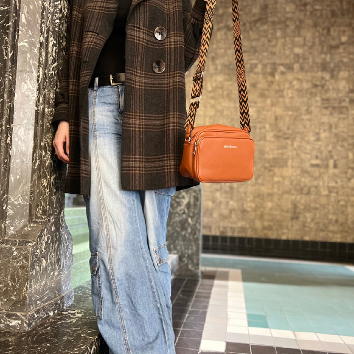 Femme en manteau à carreaux et jean large, tenant un sac à bandoulière orange Balenciaga, posant devant un mur texturé. Mode urbaine élégante.