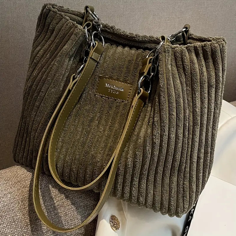 Sac à main en velours côtelé vert olive avec bandoulières en cuir, étiquette "Meichihola", posé sur un canapé beige. Accessoire mode élégant.
