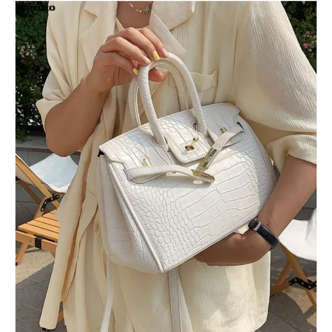 Sac à main blanc en cuir texturé croco, porté par une personne en tenue beige élégante, avec fermoir doré et poignée robuste, style chic et tendance.