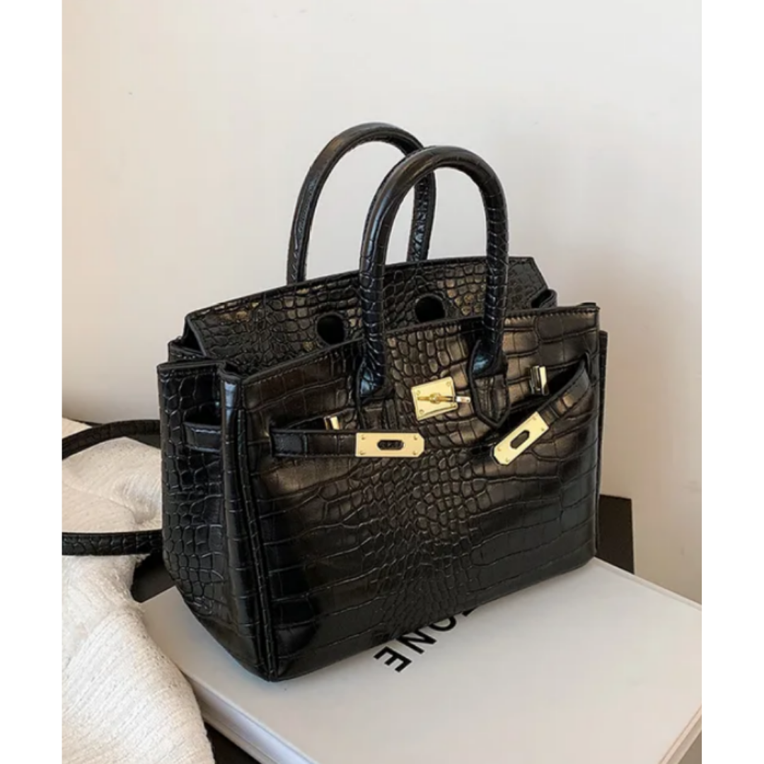 Sac à main noir en cuir croco, style Birkin, avec fermoirs dorés, posé sur un livre blanc. Accessoire mode élégant et luxueux.