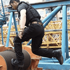 Homme en tenue tactique noire sautant sur un chantier de construction, portant des bottes robustes, avec des structures métalliques en arrière-plan.
