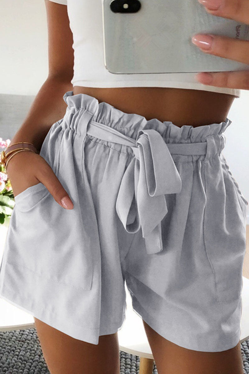 Femme portant un short taille haute gris avec ceinture nouée, top blanc court, tenant un smartphone. Mode estivale décontractée, tendance et élégante.