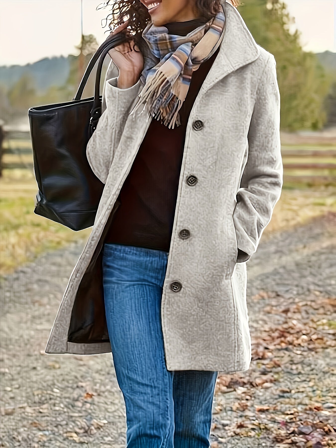Femme souriante en manteau beige, écharpe à carreaux, jeans, portant un sac noir, marchant sur un chemin rural automnal. Mode automne, style décontracté.