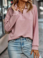 Femme portant un pull rose texturé à col zippé et un jean bleu clair, accessoirisée d'un collier doré et d'un sac à main beige. Mode décontractée.