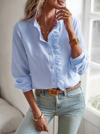 Femme portant une chemise bleue à volants, montre dorée et jean clair, posant près d'une fenêtre. Mode féminine élégante et décontractée.