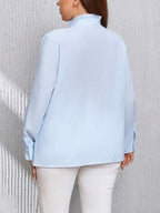 Femme portant une chemise bleue à col montant, manches longues, dos visible, sur fond de mur texturé gris. Mode féminine élégante et décontractée.