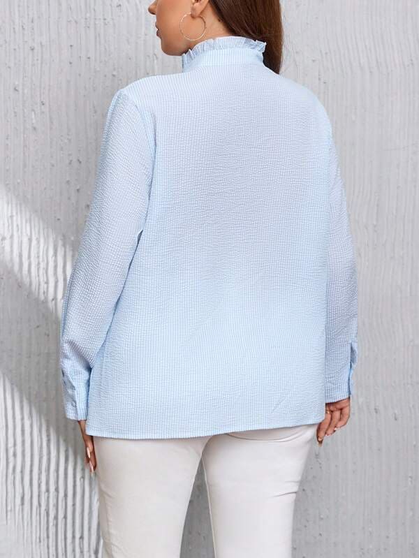 Femme portant une chemise bleue à col montant, manches longues, dos visible, sur fond de mur texturé gris. Mode féminine élégante et décontractée.