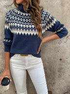 Femme portant un pull bleu à motifs nordiques, manches longues, col roulé, avec un pantalon blanc, tenant une tasse de café. Mode automne-hiver tendance.