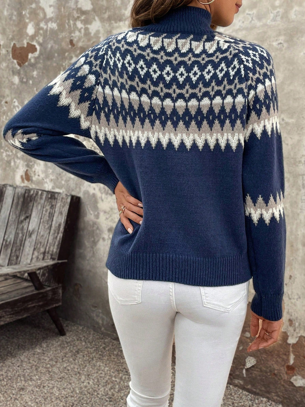 Femme portant un pull bleu à motifs nordiques blancs, manches longues, col montant, associée à un jean blanc, posant devant un mur texturé.