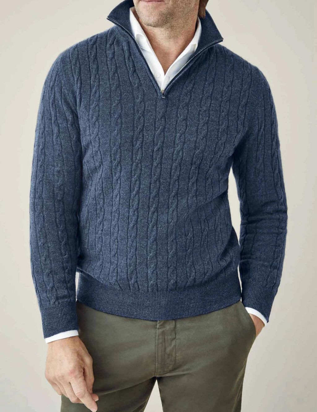 Homme portant un pull bleu à col zippé en maille torsadée, chemise blanche en dessous, pantalon vert olive. Mode masculine élégante et décontractée.
