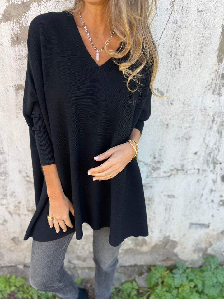 Femme portant un pull noir ample, col en V, avec des bijoux dorés, posant devant un mur texturé. Mode automne, style décontracté chic.