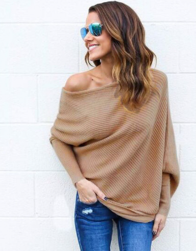 Femme souriante portant un pull oversize beige, lunettes de soleil miroir, jeans déchirés, posant contre un mur blanc. Mode décontractée chic.