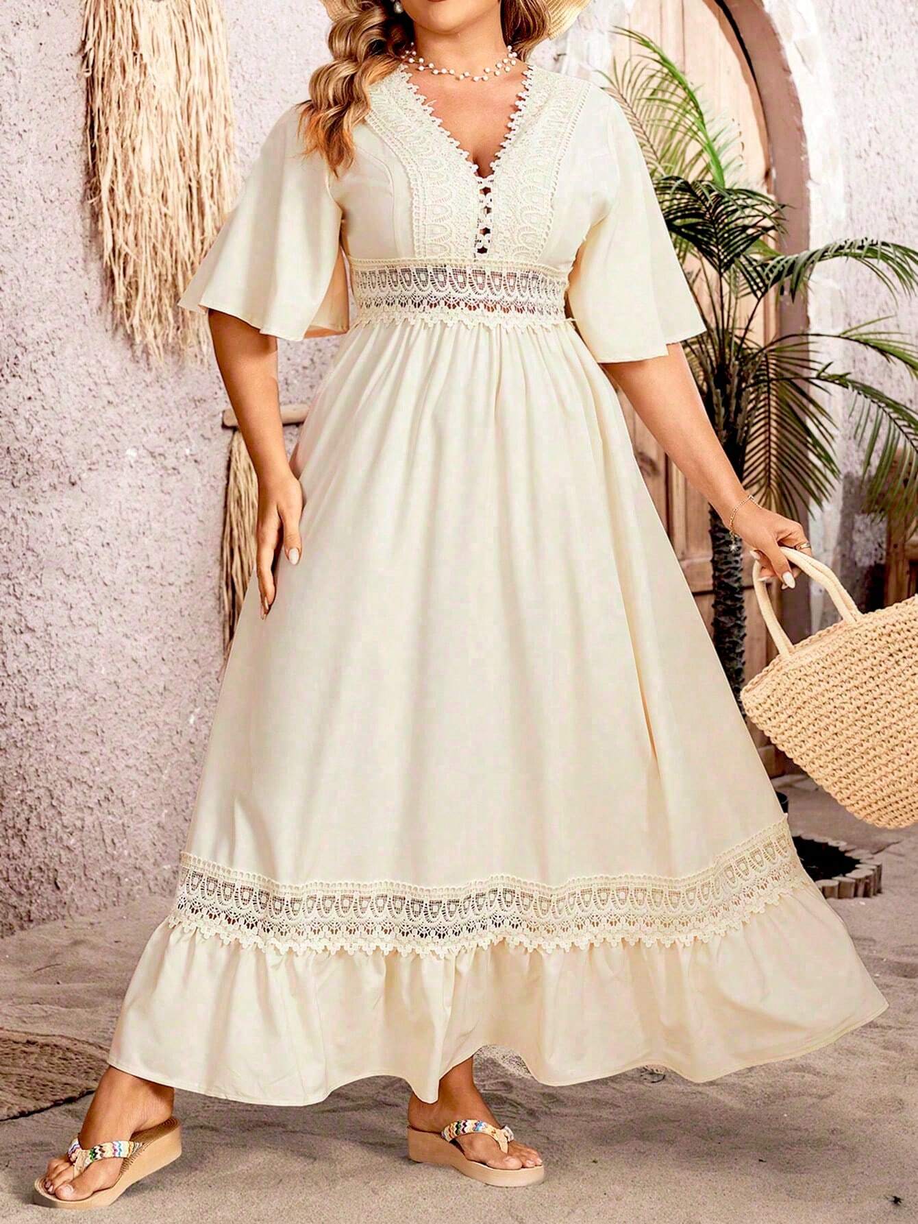 Robe longue bohème chic en dentelle blanche, manches courtes évasées, portée par une femme avec un sac en paille, devant un mur texturé.