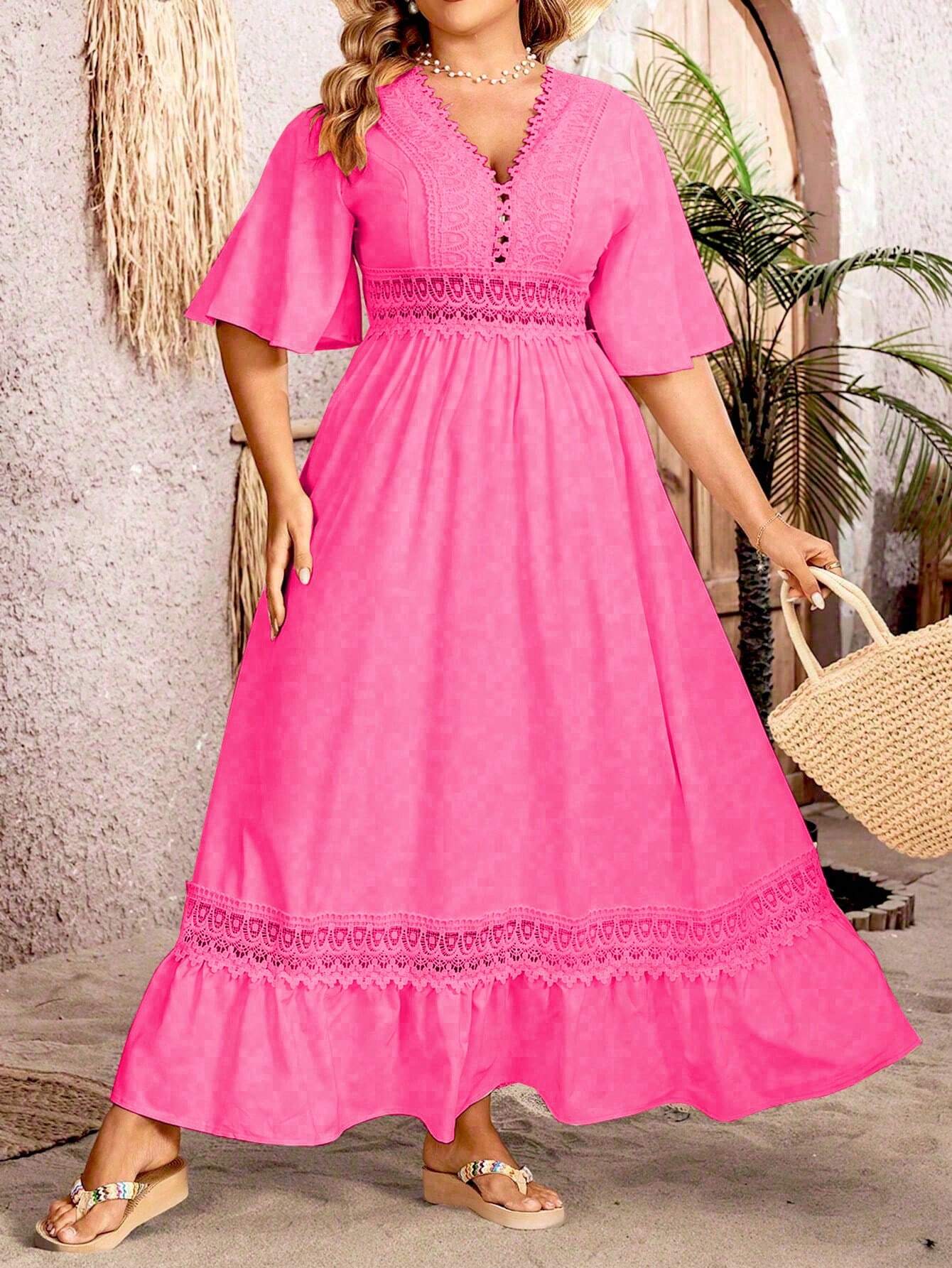 Robe longue rose vif avec dentelle, manches courtes évasées, portée par une femme tenant un sac en paille, devant un mur texturé et des plantes.