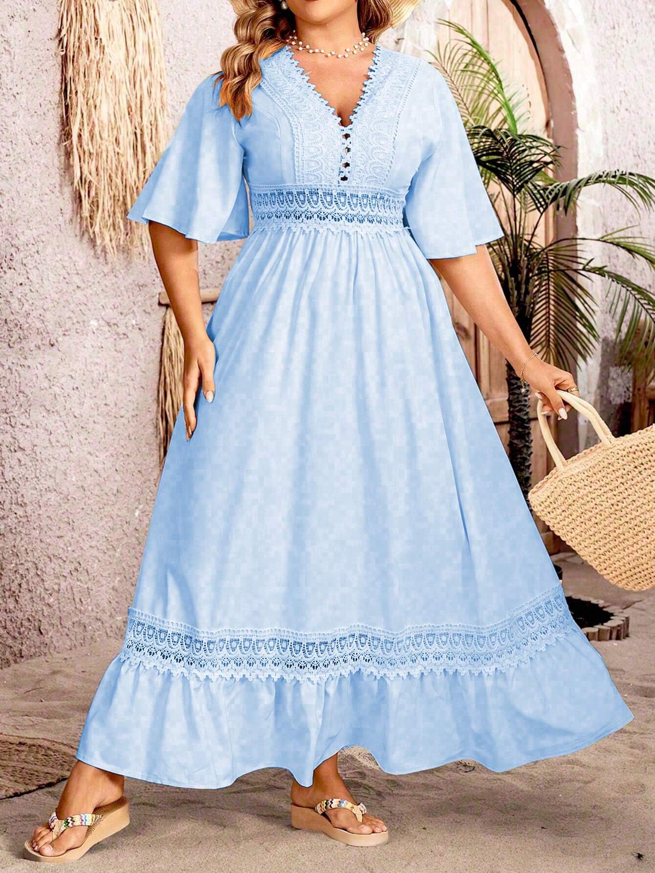 Robe longue bleue en dentelle avec manches évasées, portée par une femme tenant un sac en paille, devant un mur texturé et des plantes tropicales.