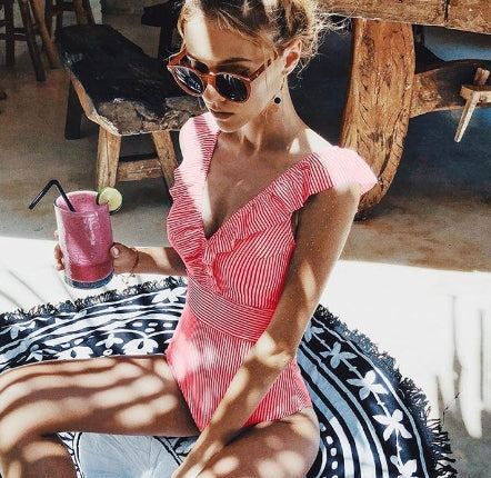 Femme en maillot de bain rayé rose, lunettes de soleil, sirotant un smoothie rose sur une terrasse ensoleillée. Vacances d'été, détente, boisson fraîche.