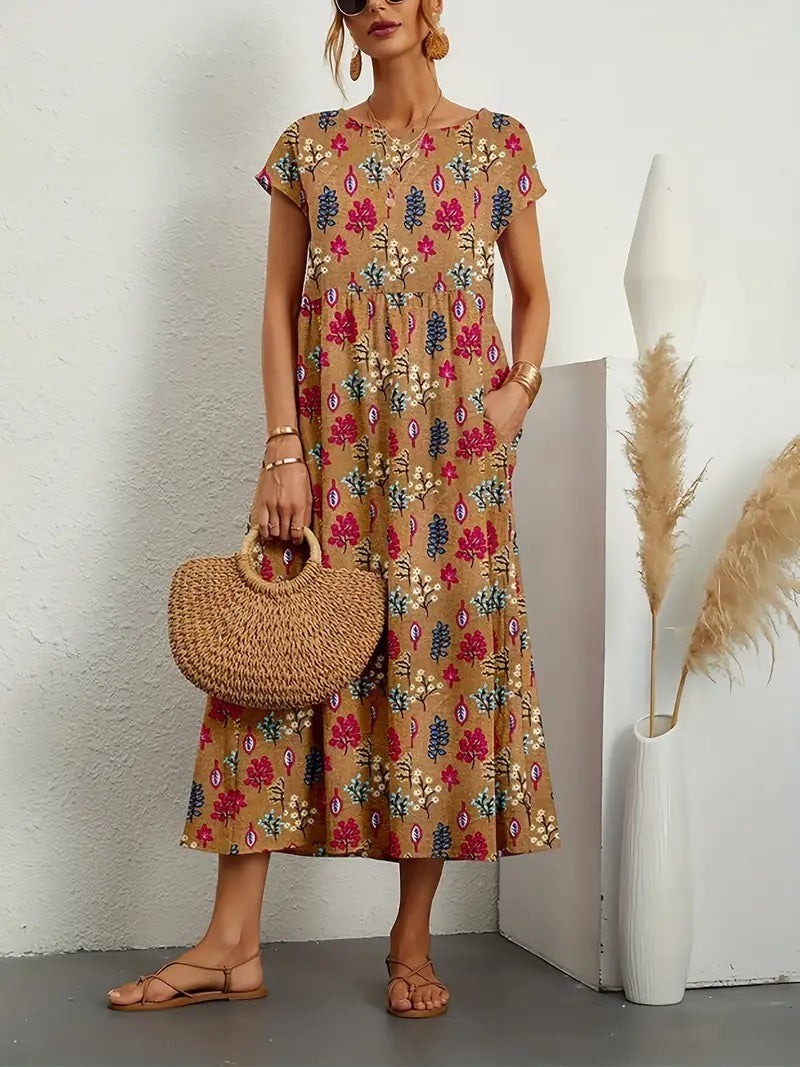 Robe longue bohème à motifs floraux colorés, femme tenant un sac en osier, sandales plates, décor minimaliste avec vase et herbes séchées.