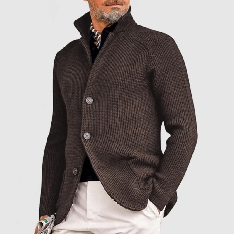 Homme portant un cardigan marron élégant avec col relevé, boutons visibles, sur chemise noire et pantalon blanc. Mode masculine chic et moderne.