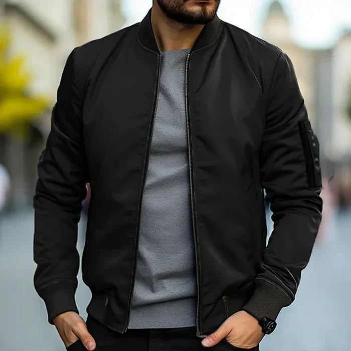 Homme portant un blouson aviateur noir, t-shirt gris, dans une rue urbaine floue. Mode masculine, style décontracté, tendance automne.