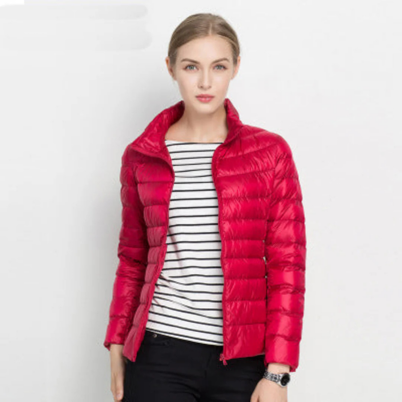 Femme portant une doudoune rouge légère, sur un fond blanc, avec un t-shirt rayé noir et blanc. Mode hiver, veste chaude, style décontracté.