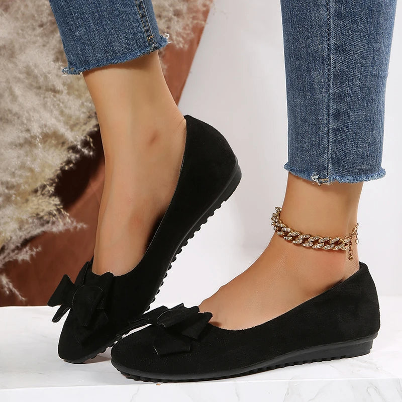 Chaussures ballerines noires en daim avec nœud, portées avec un jean bleu et bracelet doré. Mode féminine élégante et tendance.