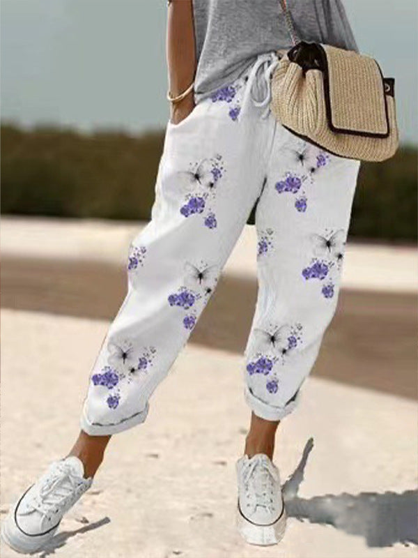 Pantalon blanc à motifs floraux violets, porté avec un t-shirt gris et des baskets blanches, sac en osier. Mode décontractée féminine estivale.