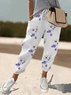 Pantalon blanc à motifs floraux violets, porté avec un t-shirt gris et des baskets blanches, sac en osier. Mode décontractée féminine estivale.