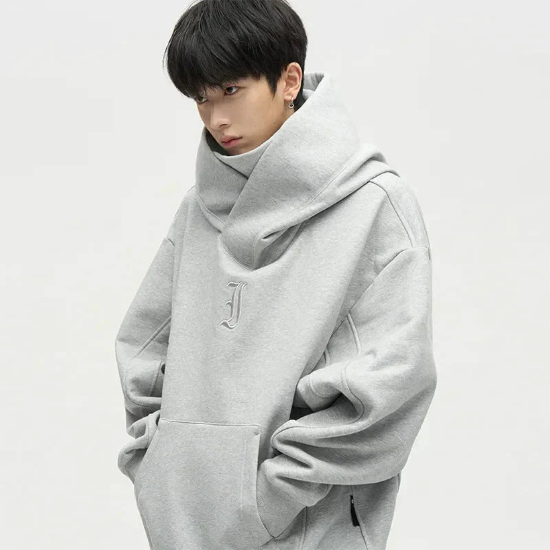 Jeune homme portant un sweat à capuche gris ample avec col croisé, mains dans les poches, style décontracté et moderne, fond blanc minimaliste.