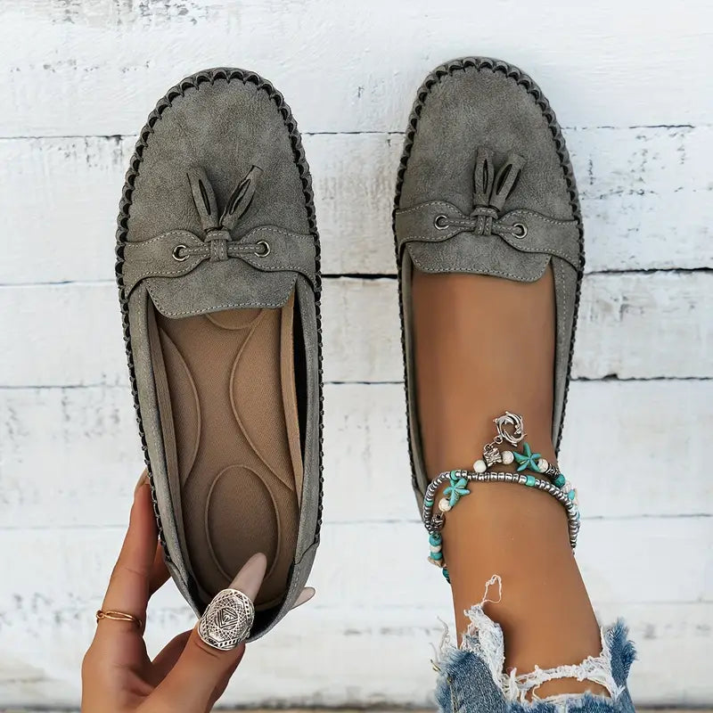 Mocassins gris en daim avec nœuds, portés par une personne avec un bracelet de cheville turquoise, sur fond de bois blanc. Mode féminine, chaussures élégantes.