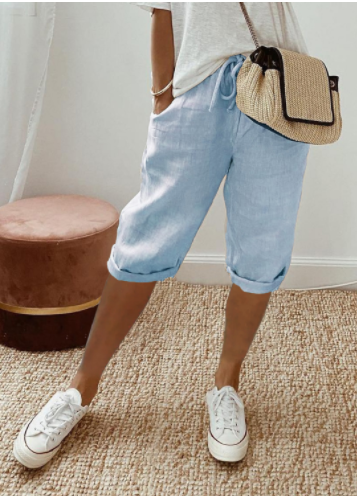 Femme portant un short en jean bleu clair, t-shirt blanc, baskets blanches et sac à main en paille, posant sur un tapis beige.