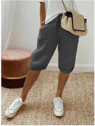 Femme portant un short gris, t-shirt blanc, baskets blanches et sac à main en paille, debout sur un tapis beige près d'un pouf rose.
